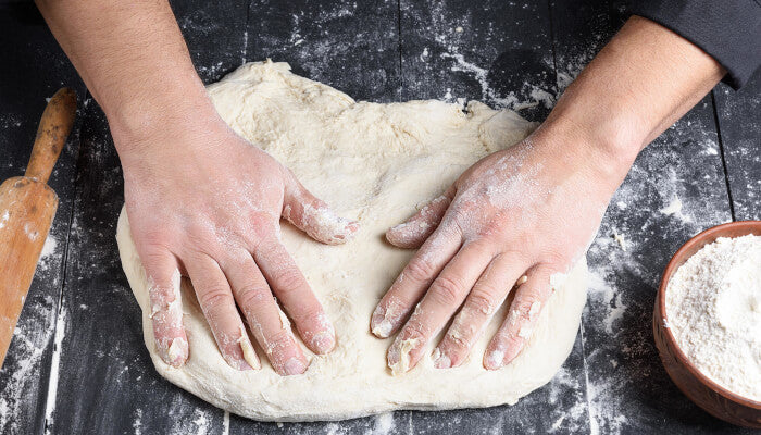 Hands pressing and stretching store-bought pizza dough on a dark floured surface with a bowl of flour in the background