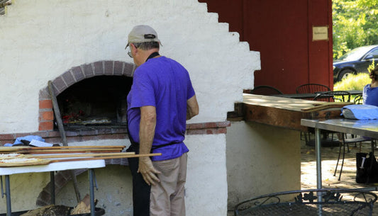 Man using a pizza peel at an outdoor brick pizza oven with a classic arched brick dome in a backyard setting