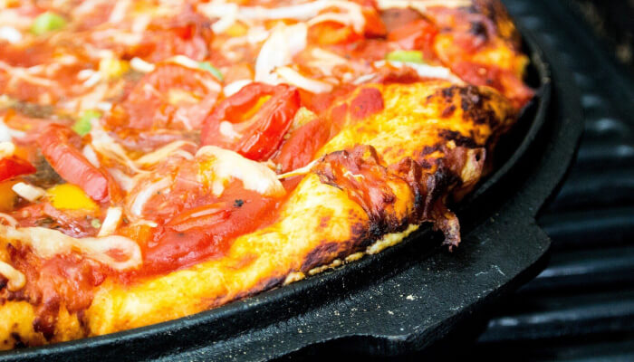 Topped pizza cooking on a ridged cast iron grill pan on a gas grill with visible char on the crust and melted cheese