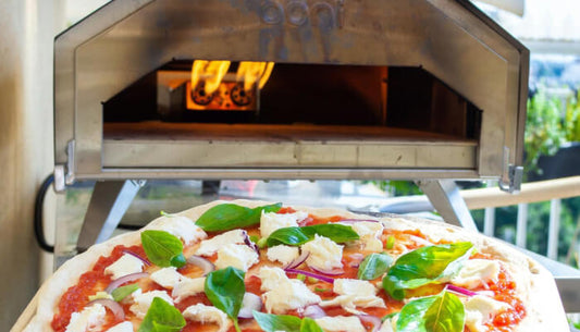 Fresh pizza with basil and mozzarella in front of a wood-fired outdoor pizza oven with visible flames inside