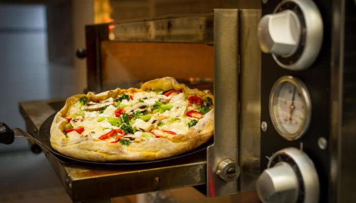 Pizza being pulled from a natural gas pizza oven on a metal peel with temperature gauges visible on the oven exterior showing a fully cooked pizza with melted cheese and fresh toppings
