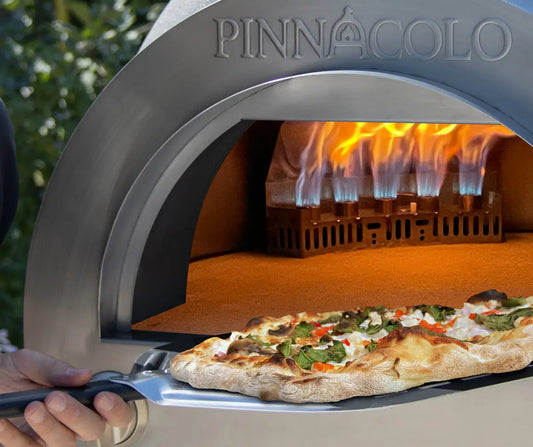 Pizza coming out of a gas-fired outdoor pizza oven on a peel with the burner flame behind it
