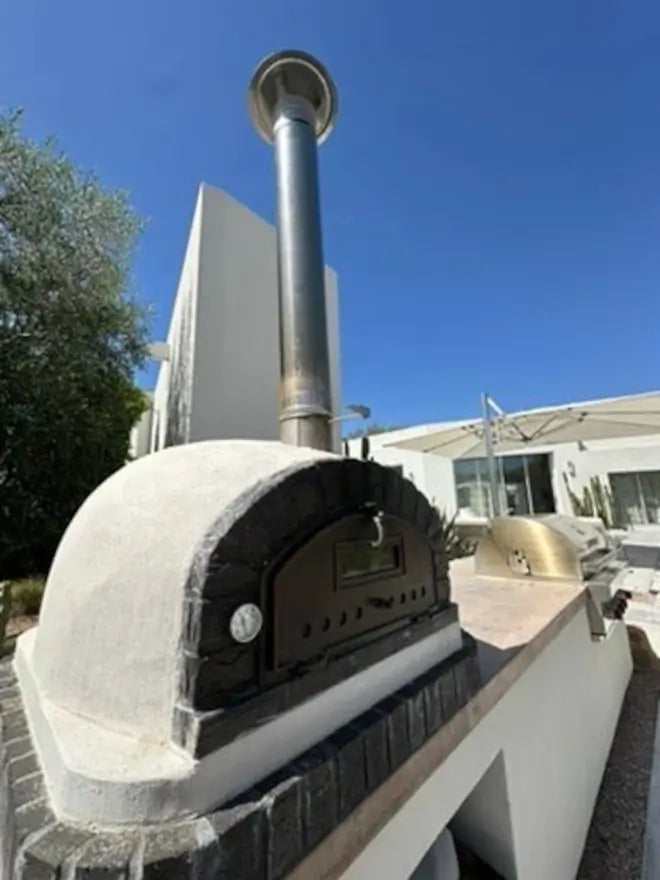 Stainless chimney cap on a tall extension above a stucco Authentic Pizza Ovens brick oven in an outdoor kitchen