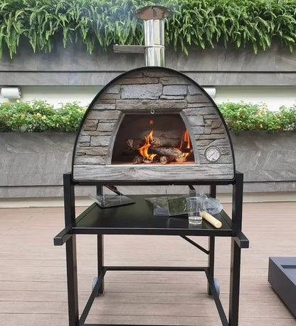 Black Maximus Prime pizza oven on the APO stand with a fire burning, fern wall behind