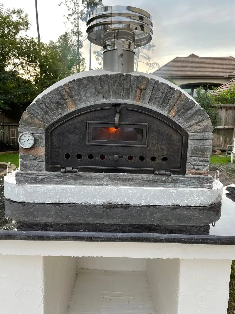 Buena Ventura Preto brick pizza oven at dusk with the fire glowing through the door window, set on a granite topped base