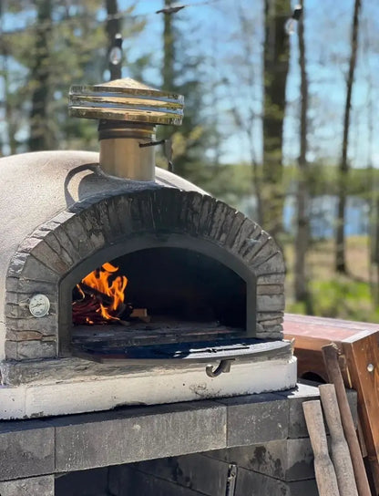 Buena Ventura Preto pizza oven with the drop down door lowered, fire burning inside, and a rain cap on the chimney