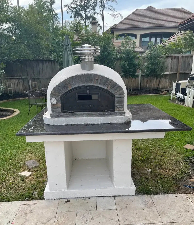 Buena Ventura Preto pizza oven with chimney extension and rain cap on a white stucco block base with a black granite top