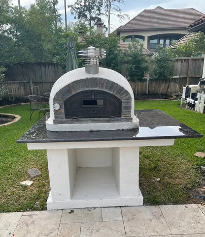 Buena Ventura Preto pizza oven with chimney extension and rain cap on a white stucco block base with a black granite top