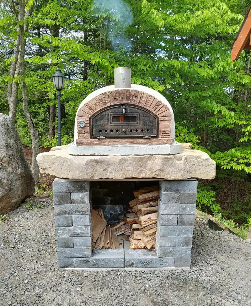 Buena Ventura Sierra pizza oven lit and smoking on a natural stone slab over a gray block base with firewood storage