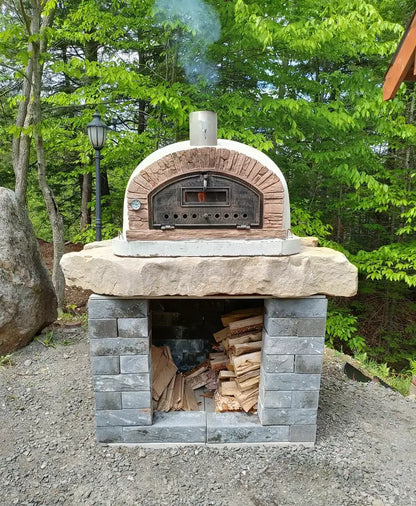 Buena Ventura Sierra pizza oven lit and smoking on a natural stone slab over a gray block base with firewood storage