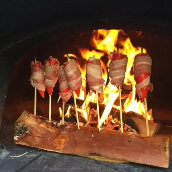 Bacon wrapped jalapeno poppers on skewers standing in a log beside the fire in a brick pizza oven