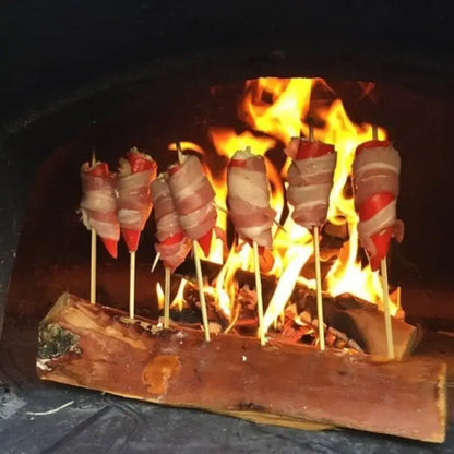Bacon wrapped jalapeno poppers on skewers standing in a log beside the fire in a brick pizza oven
