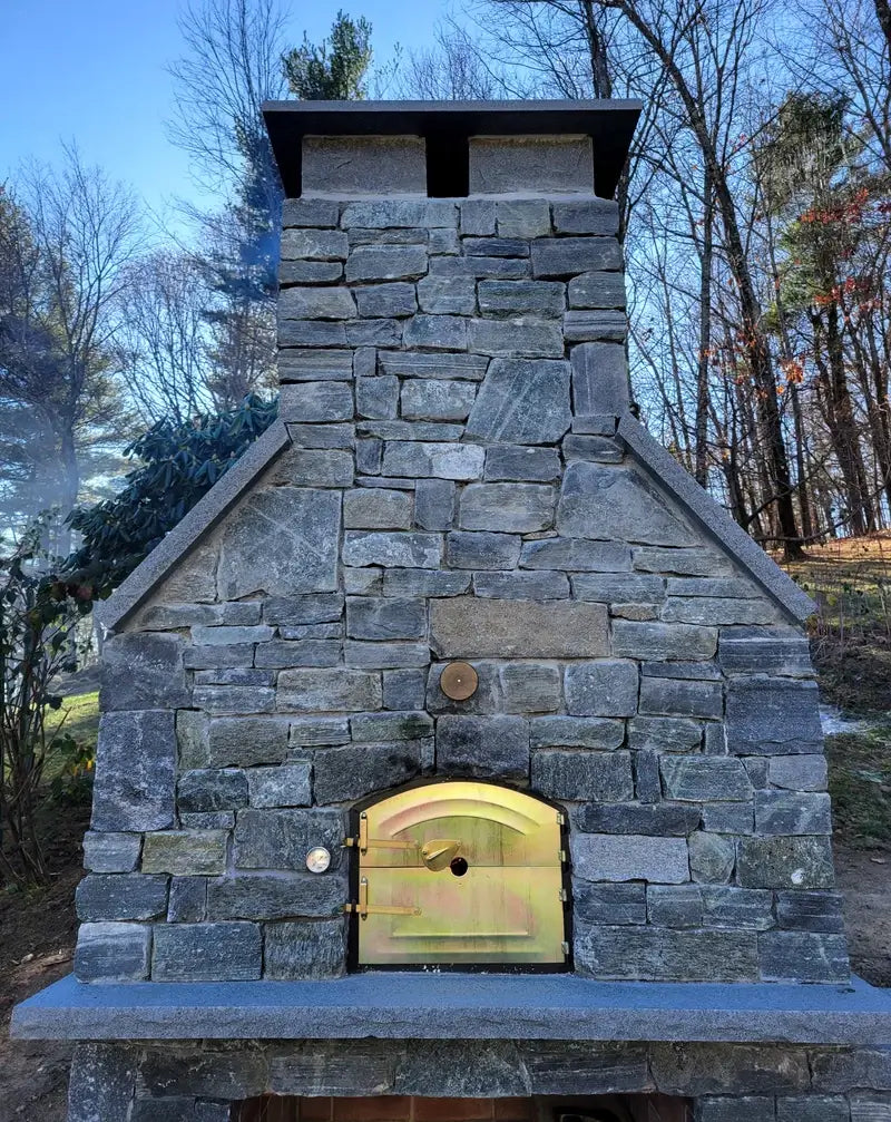 Close view of the stone clad builders custom Lisboa showing the arched brass door, the thermometer set into the stone, and the chimney rising above the shelf
