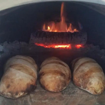 Three bread loaves baking on the floor of a brick pizza oven with a log burning at the back