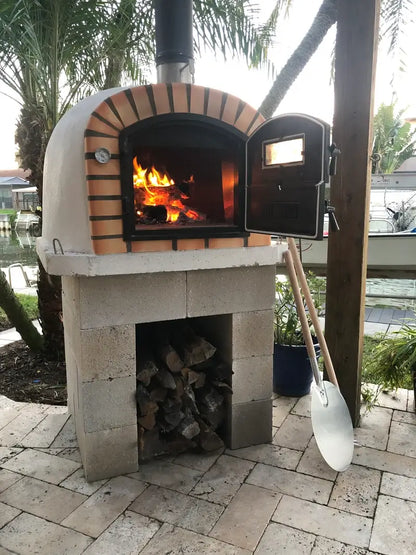 Lisboa Premium pizza oven on a concrete block base with the door open and a fire burning, wood stored below, peel and ash shovel beside it