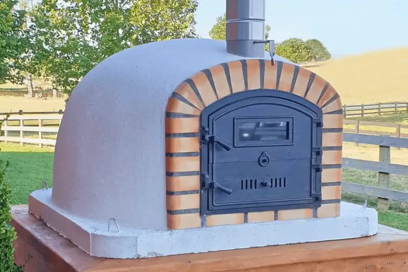 Lisboa Premium brick pizza oven installed outdoors on a masonry base with a stainless chimney extension, fields behind