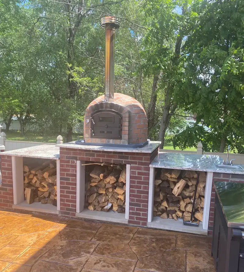 Rustic Lisboa pizza oven set on a brick base with granite counters and wood storage, tall chimney extension with rain cap