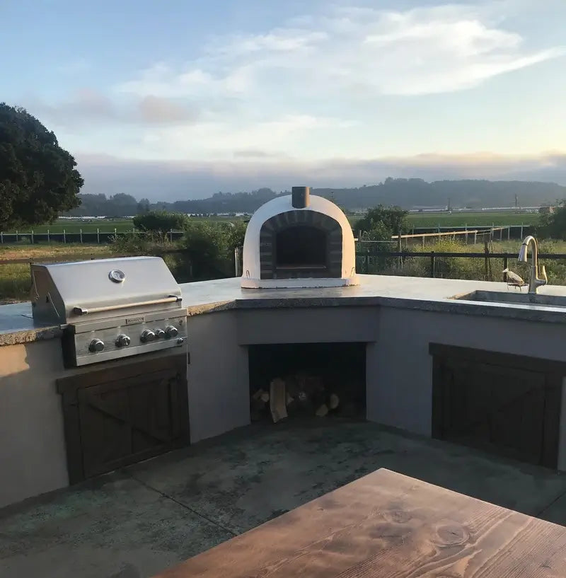 Lisboa Stone Arch pizza oven set into an outdoor kitchen counter beside a built-in grill, farmland and evening sky behind