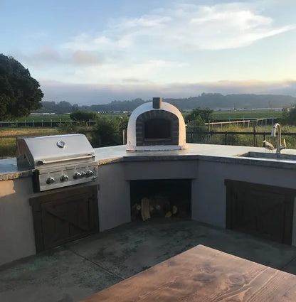 Lisboa Stone Arch pizza oven set into an outdoor kitchen counter beside a built-in grill, farmland and evening sky behind