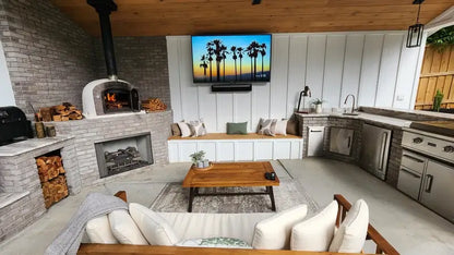 Lume Alto Premium pizza oven lit in the corner of a covered outdoor living room with a fireplace, seating, and kitchen