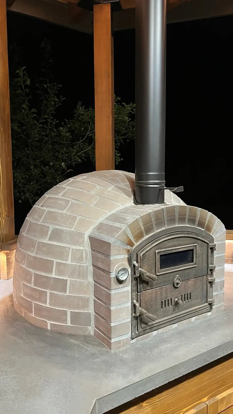 Lume Alto Tudo pizza oven at night on a concrete counter in a timber gazebo, black chimney pipe rising from the cast stone dome