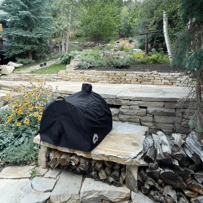 Authentic Pizza Ovens black cover over a Maximus oven on a stone bench with firewood stacked beneath it in a terraced garden