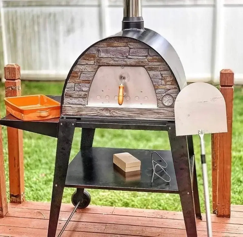 Black Maximus Mobile pizza oven on the Authentic Pizza Ovens stand on a deck with the included stainless peel and orange ceramic baking dish beside it