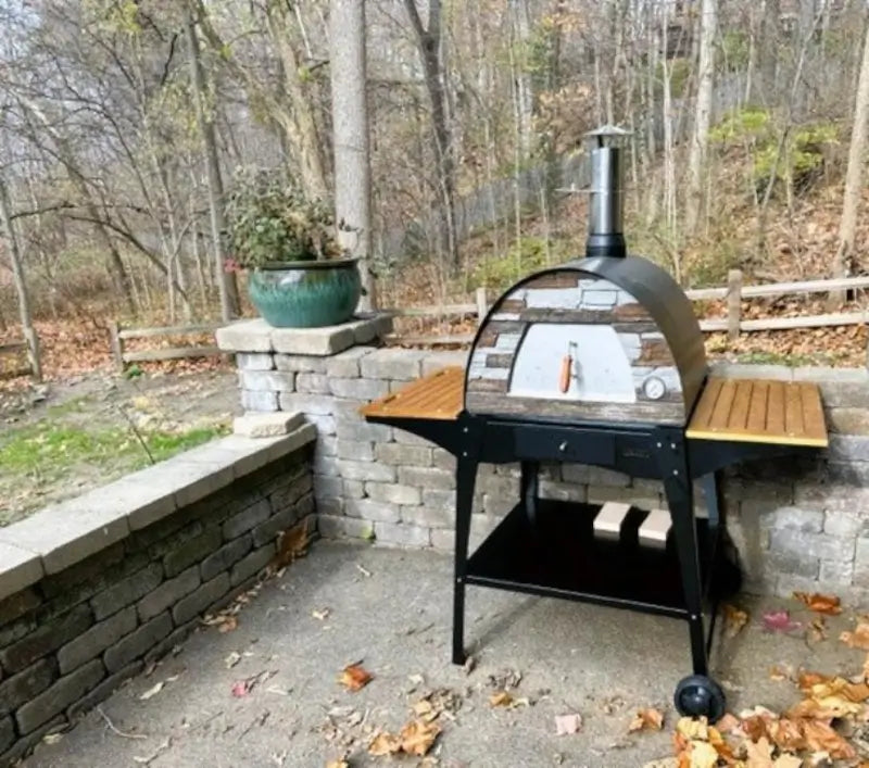 Black Maximus Mobile pizza oven on the Authentic Pizza Ovens stand with wooden side shelves on a stone walled patio in autumn