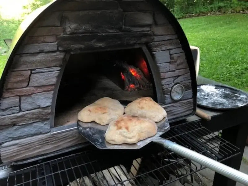 Three bread loaves on a peel at the mouth of a black Maximus Mobile pizza oven with coals glowing inside