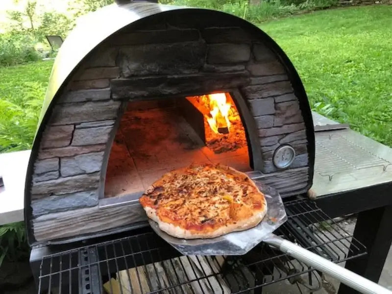 Pizza on a stainless peel coming out of a black Maximus Mobile pizza oven with the fire burning at the back of the tile floor