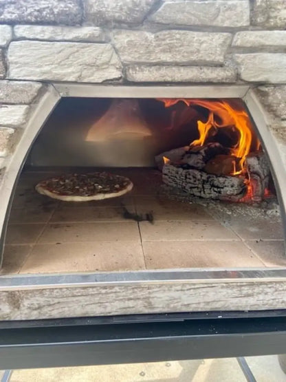 Inside the Maximus Mobile pizza oven, a pizza on the tile floor with a log burning at the back of the stainless chamber