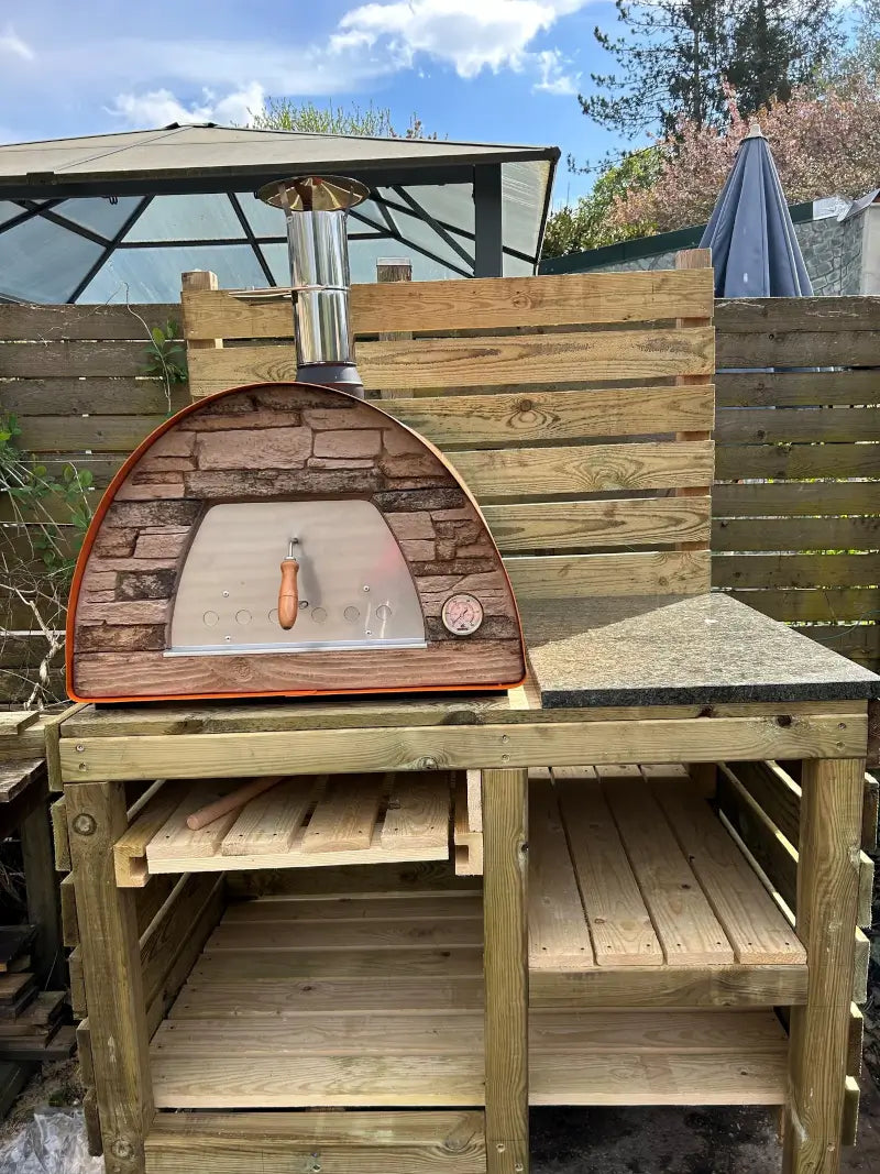 Orange Maximus Mobile pizza oven on a homemade timber bench with a granite landing beside it under a garden gazebo