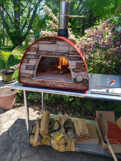 Red Maximus Mobile pizza oven with the fire lit on a stainless prep table in a garden, bagged firewood and the ceramic dish on the shelf below