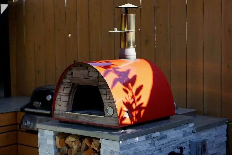 Red Maximus Mobile pizza oven with the door off on a stacked stone base with a firewood bay, evening light across the red body