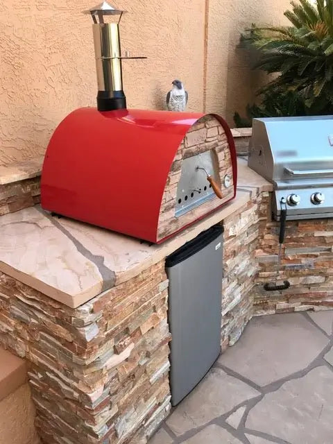 Red Maximus Mobile pizza oven built into a stacked stone outdoor kitchen counter beside a grill and a fridge