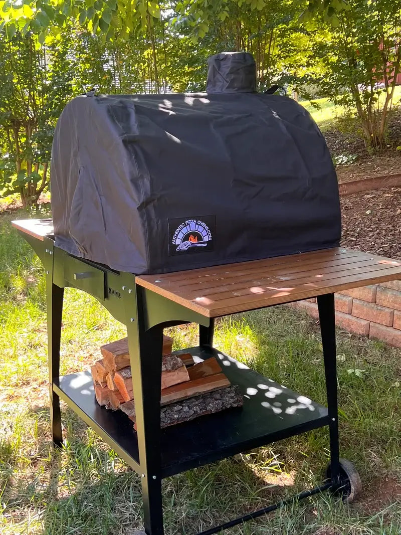 Maximus Mobile pizza oven under its cover on the Woody stand, firewood stacked on the lower shelf and a wood slat side shelf extended