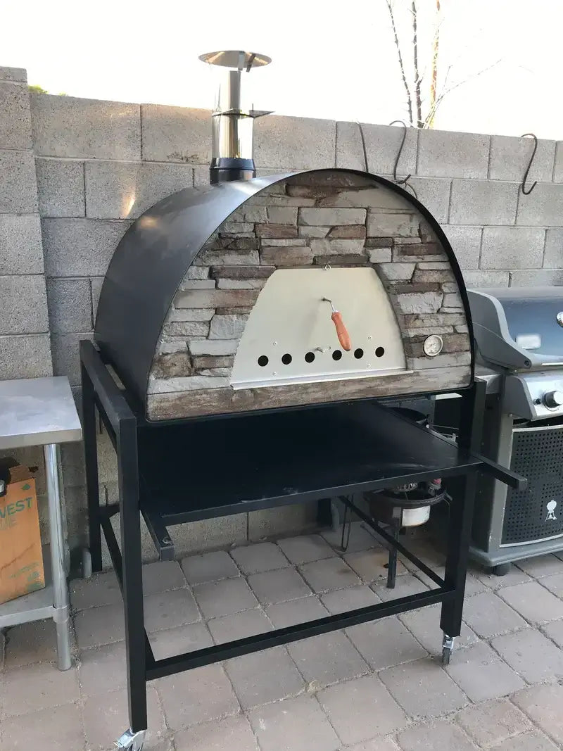 Black Maximus Prime pizza oven on the APO rolling cart against a block wall on a paver patio