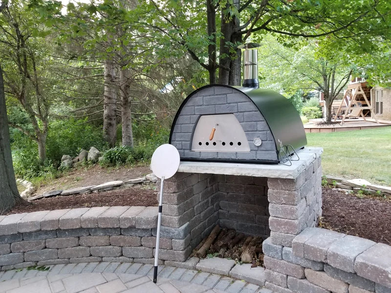 Black Maximus Prime pizza oven on a paver block base with a wood bay below and a round peel leaning beside it, trees behind
