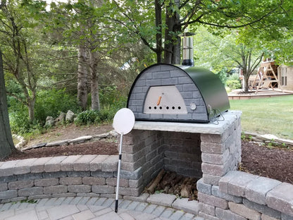 Black Maximus Prime pizza oven on a paver block base with a wood bay below and a round peel leaning beside it, trees behind
