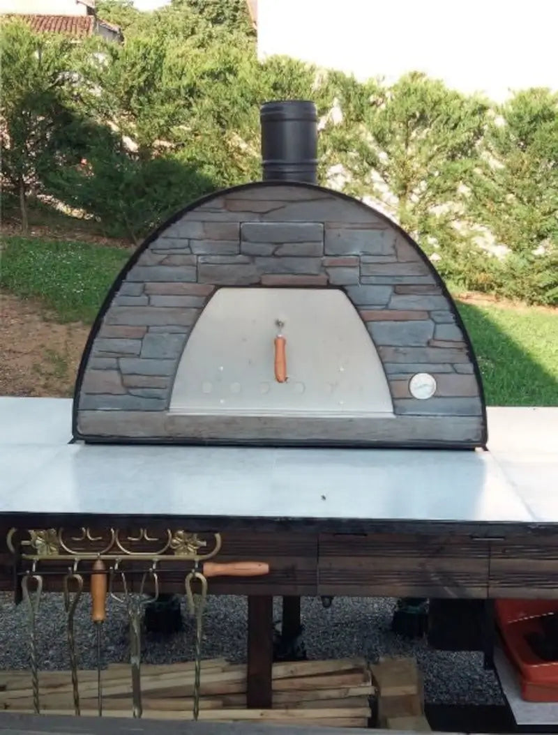 Black Maximus Prime pizza oven on a white tiled outdoor counter with pizza tools hung on hooks below