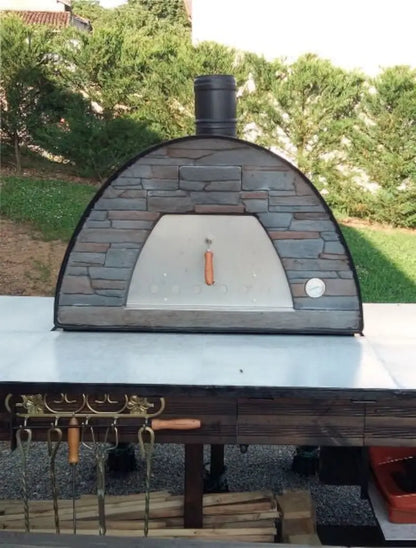 Black Maximus Prime pizza oven on a white tiled outdoor counter with pizza tools hung on hooks below