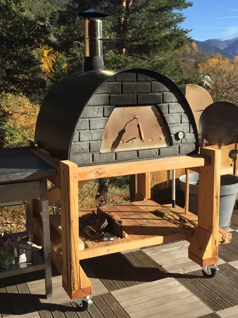 Black Maximus Prime pizza oven on a homemade timber cart with casters on a deck, autumn trees and mountains behind