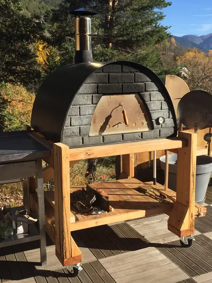Black Maximus Prime pizza oven on a homemade timber cart with casters on a deck, autumn trees and mountains behind
