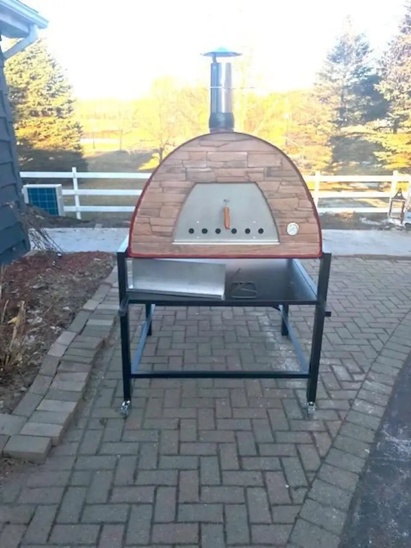 Red Maximus Prime pizza oven on a black rolling cart with the included peel on the shelf, on a brick driveway in winter