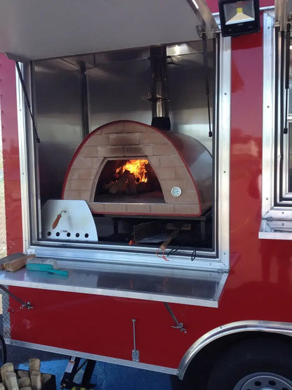 Maximus Prime pizza oven with the fire lit inside a red food trailer, the stainless door and brush on the serving counter