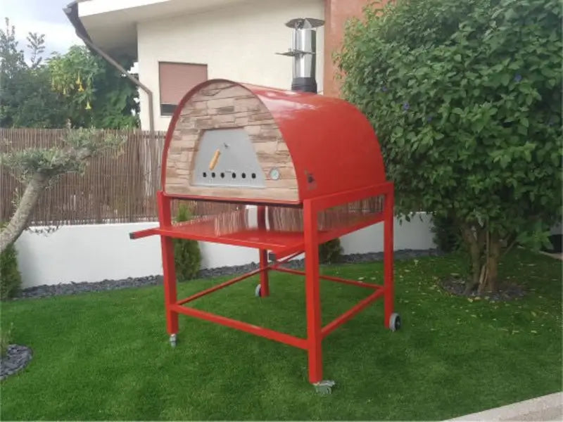 Red Maximus Prime pizza oven on a matching red steel cart with a side shelf, parked on a lawn beside a garden