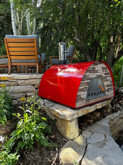 Red Maximus Prime pizza oven on a stone wall base with firewood stored below, garden chairs and aspens behind