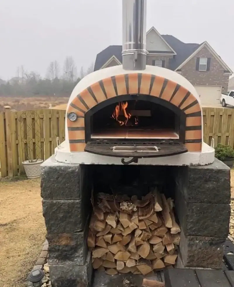Authentic Pizza Ovens Pizzaioli Premium with the door down and a fire burning, installed on a stacked block base with firewood below