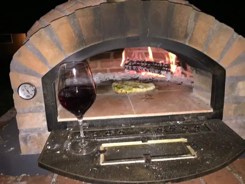 Rustic Pizzaioli at night with the door down, a pizza baking beside the fire, and a glass of red wine resting on the open door