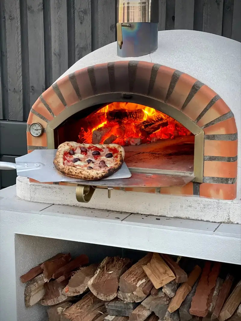 Pizza on an aluminum peel at the door of an Authentic Pizza Ovens Pizzaioli Standard, fire at the back, firewood stacked in the base